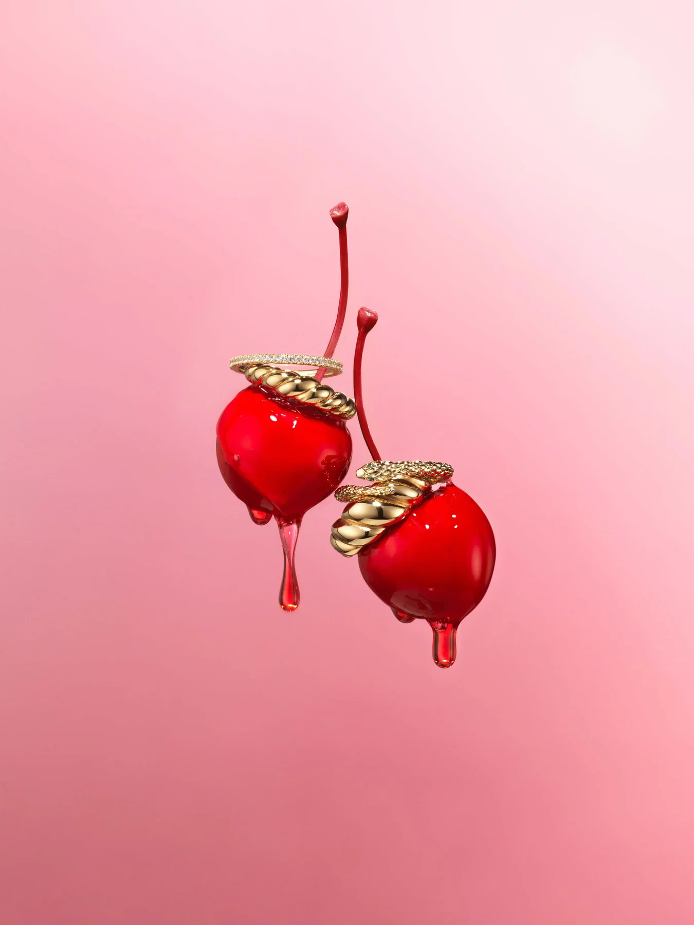 Two glossy red cherries capped with gold, dripping, on pink