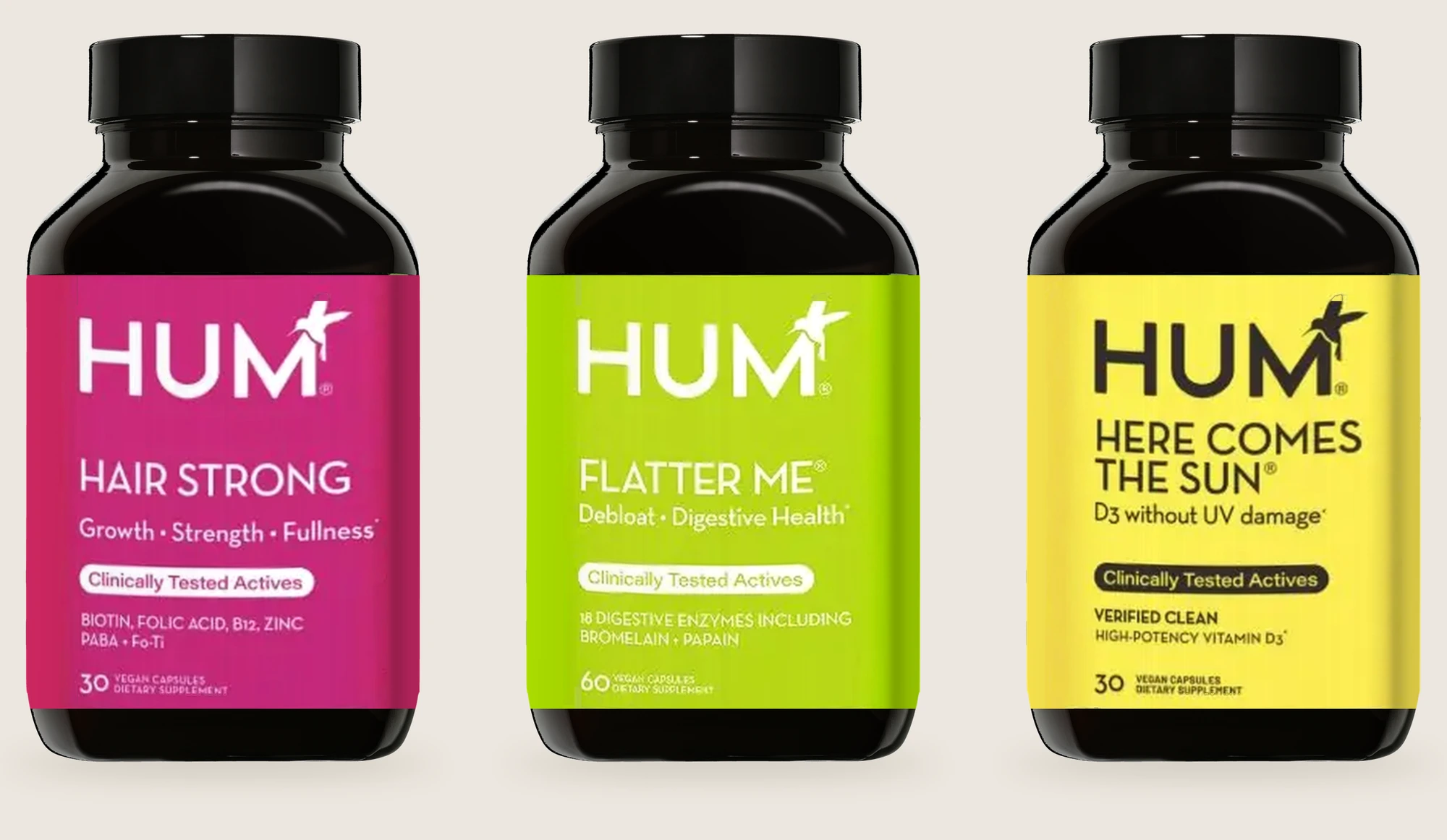 Previous HUM packaging shown straight-on: three identical black bottles on a warm stone backdrop with Hair Strong in magenta, Flatter Me in lime and Here Comes the Sun in yellow, each with the HUM wordmark and attached bird at the top of the label