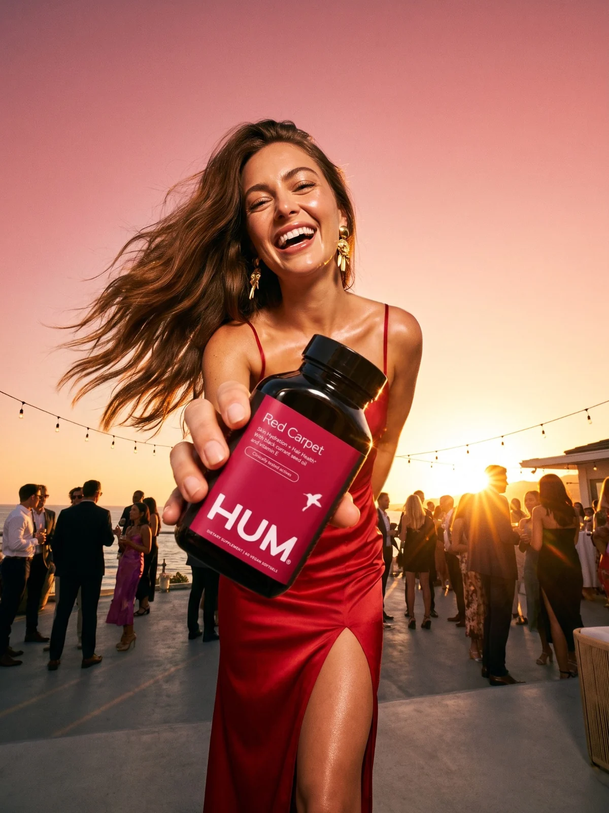 Woman in a red dress laughing at a sunset terrace party, holding out a Red Carpet bottle